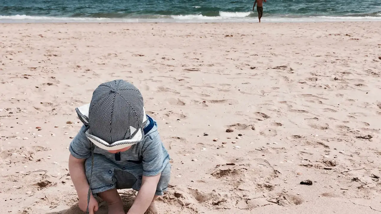 Kleines Kind mit Hut spielt im Sand am Strand der Algarve. Im Hintergrund läuft ein Mann ins Meer. Ein perfekter Tag für Algarve mit Kindern.