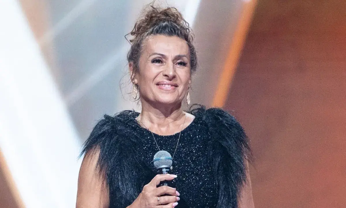 Uśmiechnięta finalistka, która mogła wygrać Voice Senior, w czarnej, błyszczącej sukni z futrzanym kołnierzem, trzyma mikrofon.