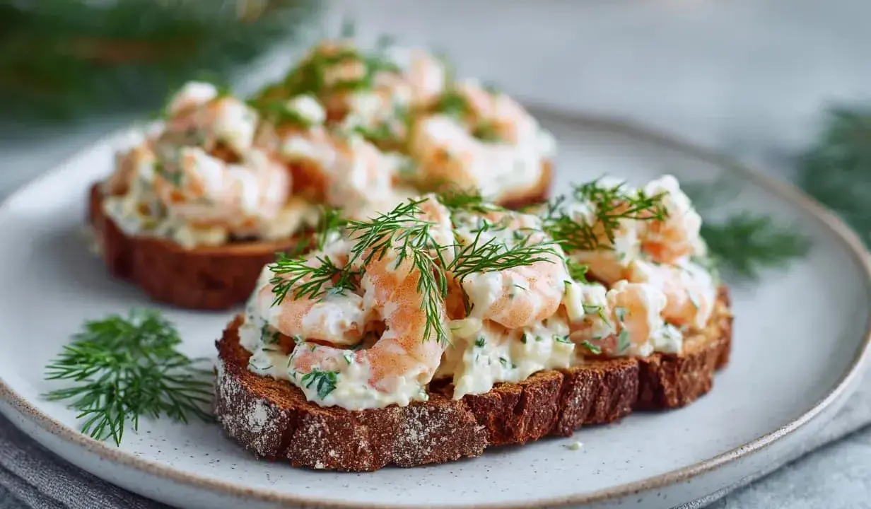 Två toast Skagen med dill och räkor på mörkt bröd, serverade på en tallrik.