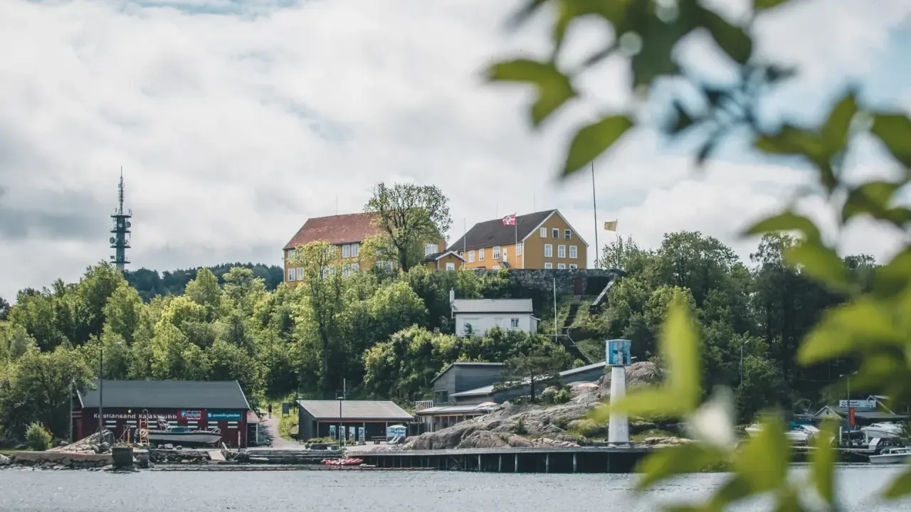 Widok na wybrzeże w Kristiansand, Norwegia. Żółte budynki na wzgórzu, czerwony klub kajakowy i wieża telekomunikacyjna.
