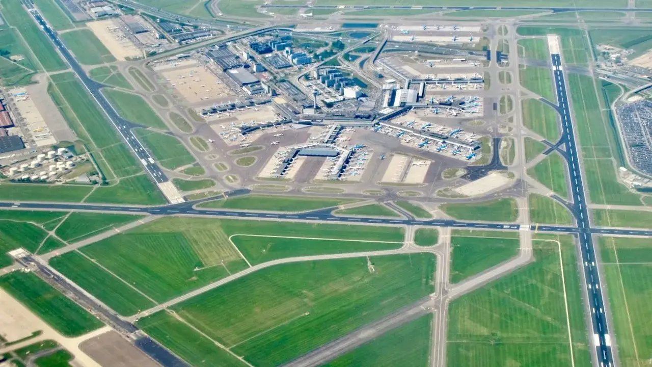 Ögonblicksbild över ams flygplats med flygplan på marken och i luften, omgivet av gröna fält och vägar.