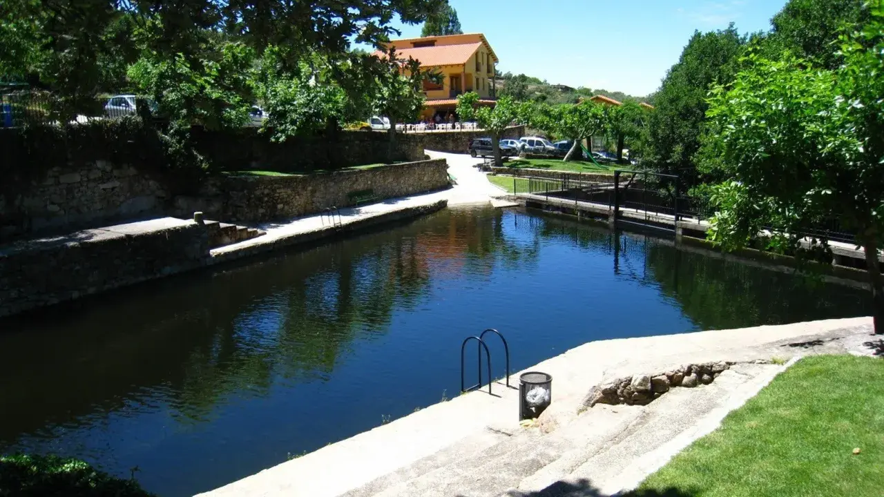 La piscina natural de Casas del Monte, rodeada de vegetación y con un edificio al fondo, invita a un refrescante baño.