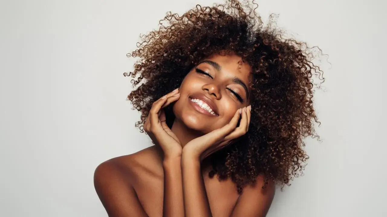 Una mujer sonríe con los ojos cerrados, su cabello rizado enmarca su rostro. Su piel radiante y su expresión serena sugieren cómo parecer más joven.