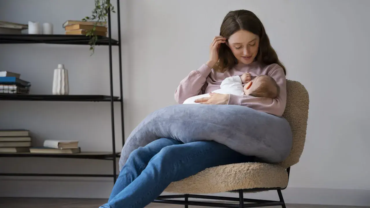 Szukasz idealny fotel do karmienia? Młoda mama z dzieckiem na rękach, otulona poduszką, siedzi na wygodnym, pluszowym fotelu.