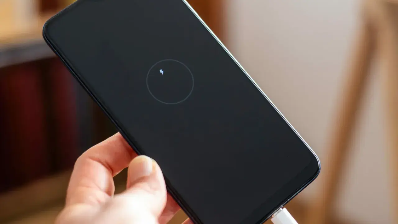 Dłoń trzyma smartfon z ikoną ładowania zwrotnego na czarnym ekranie. Podłączony biały kabel USB.