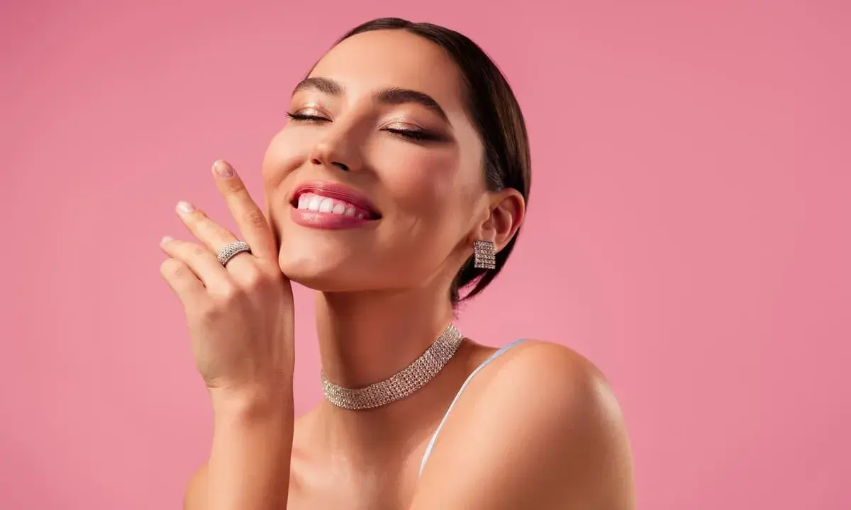 Mujer sonríe con ojos cerrados, luciendo un maquillaje romántico, joyas brillantes y un fondo rosa.