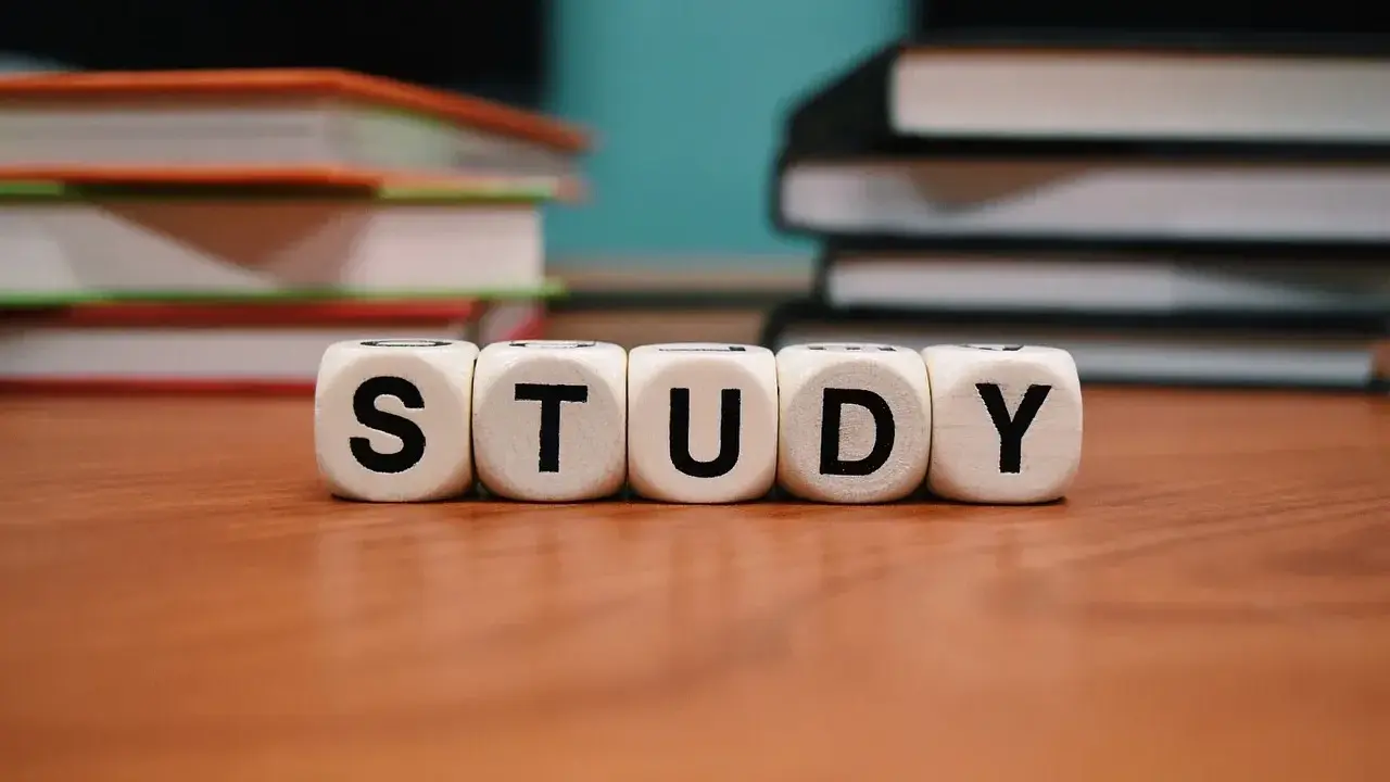 Białe kostki z literami "STUDY" na drewnianym stole, w tle stosy książek.