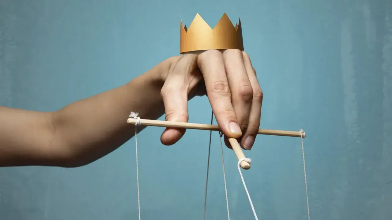 Dłoń z papierową koroną na palcach, sterująca marionetką. Symbol władzy i despoty.