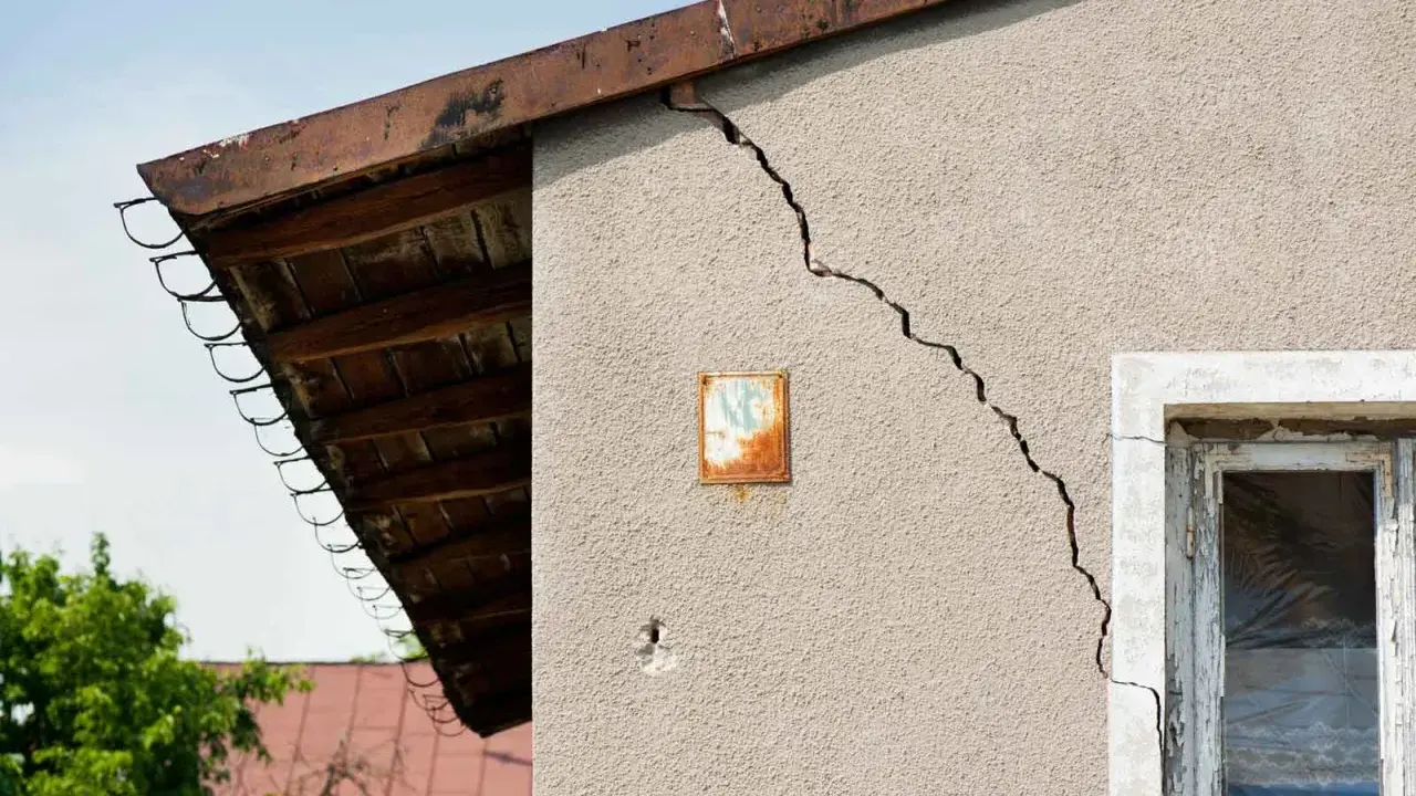 Grieta en pared de edificio, indicando problemas de **seguridad estructural**. Se observa un alero de madera y un cielo azul.