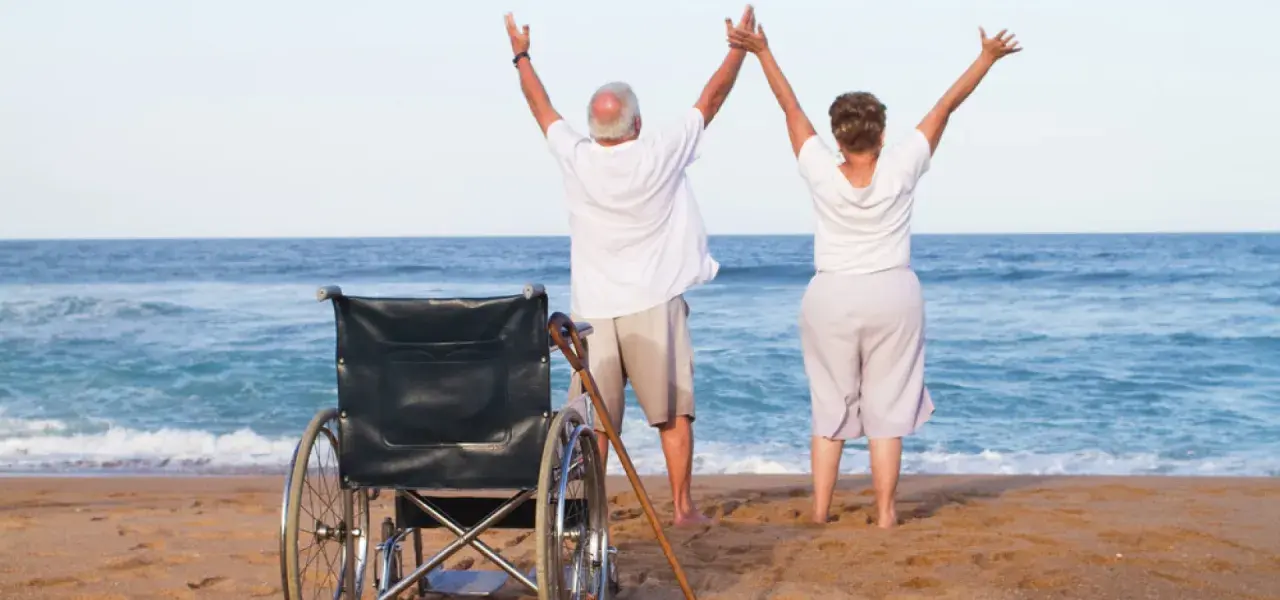Szczęśliwi seniorzy cieszą się urlopem. Wózek inwalidzki i laska na plaży świadczą o tym, że to udany turnus rehabilitacyjny nad morzem.