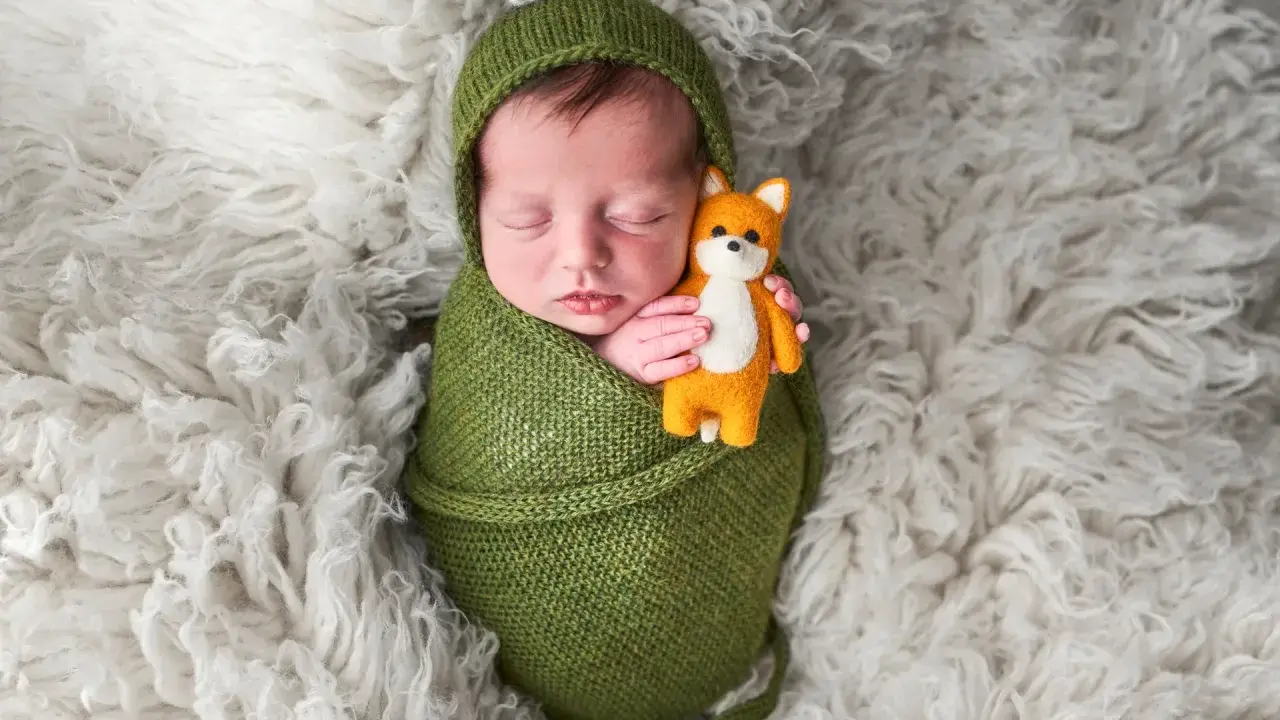 Dolci immagini neonati: un bimbo avvolto in una coperta verde tiene stretto un piccolo volpino di peluche, dormendo sereno su un soffice tappeto bianco.