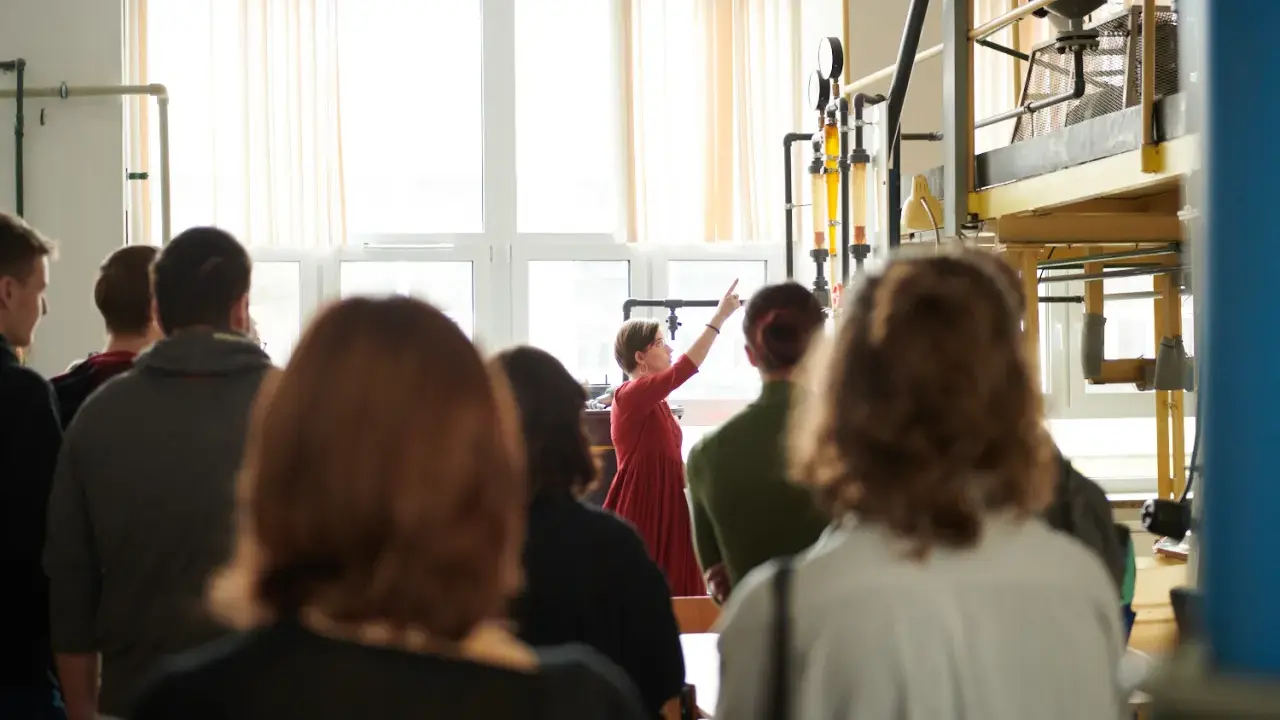 Grupa studentów na zajęciach praktycznych. Kobieta w czerwonej sukience tłumaczy coś przy aparaturze. To może być część ich studiów magisterskich zaocznych w Warszawie.