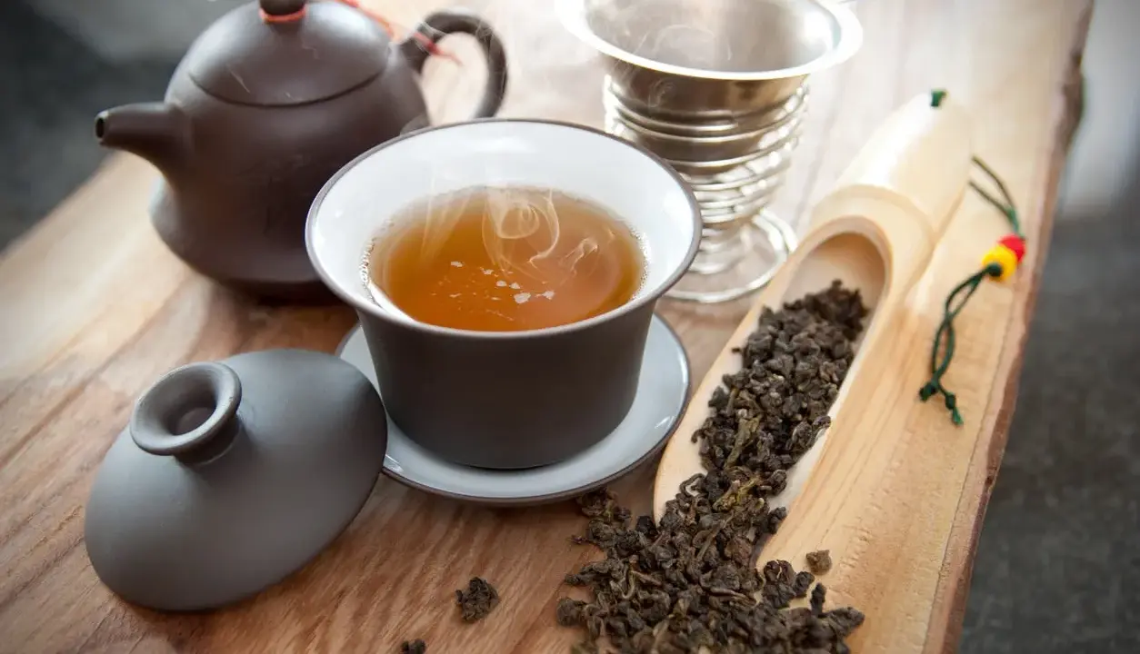 Dampfender Oolong Tee in einer Tasse, daneben Teekanne, Sieb und lose Teeblätter. Die Oolong Tee Wirkung kann belebend sein.