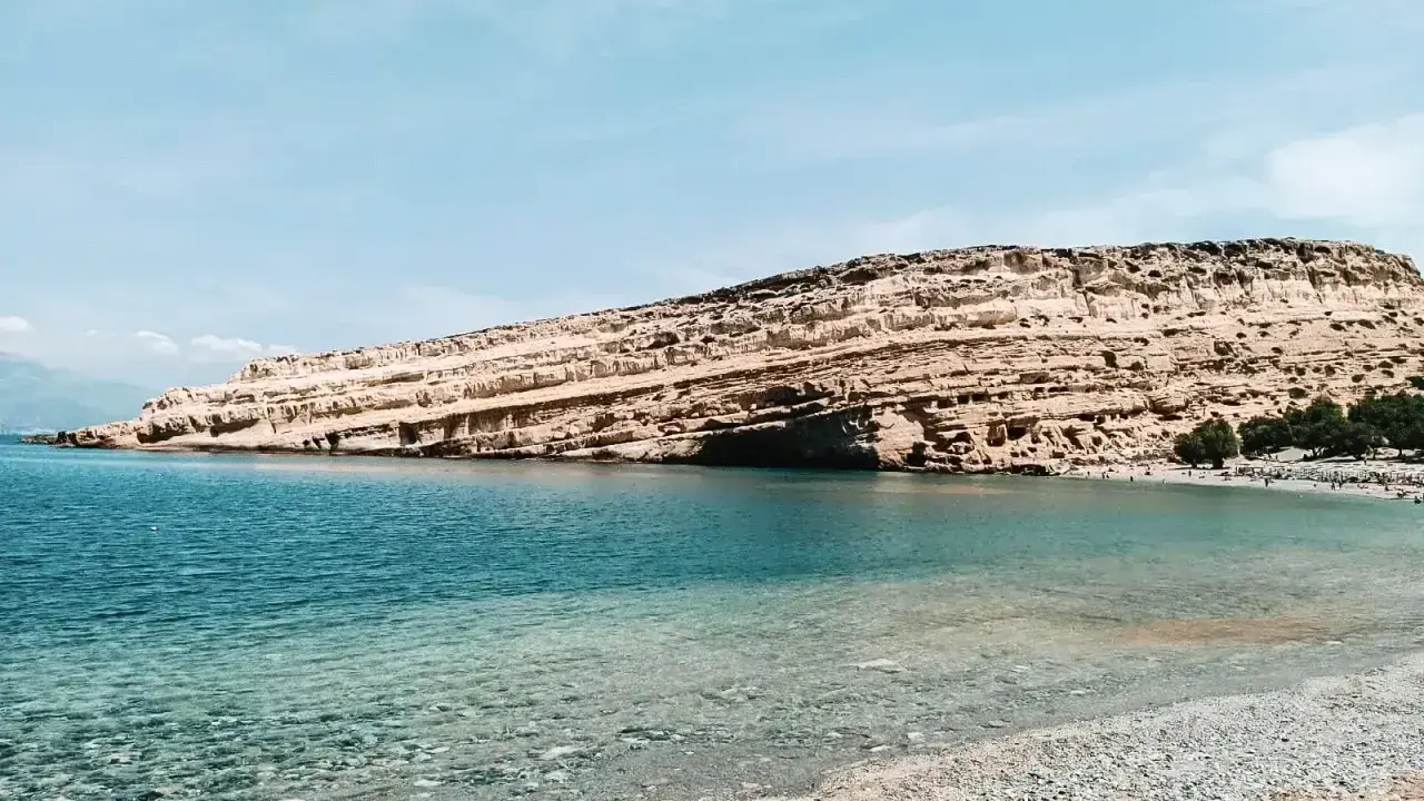 Plaża z kamienistym brzegiem i skalistym klifem w tle, nad turkusowym morzem.