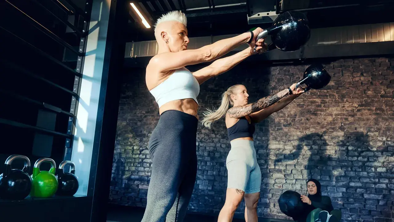 Ein Paar trainiert mit Kettlebells im Fitnessstudio. Sie heben Gewichte, während eine dritte Person im Hintergrund sitzt.