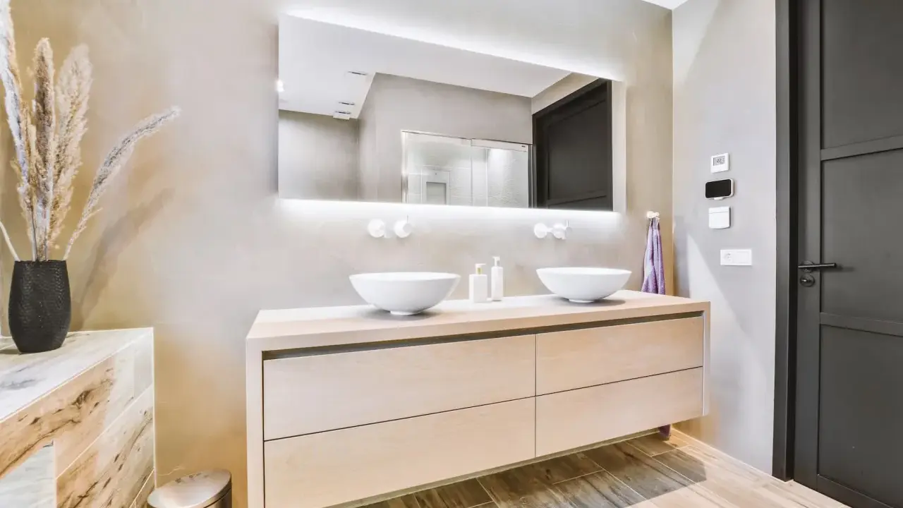 Elegantes baños modernos pequeños con lavabos dobles sobre mueble de madera clara, espejo iluminado y pared texturizada.