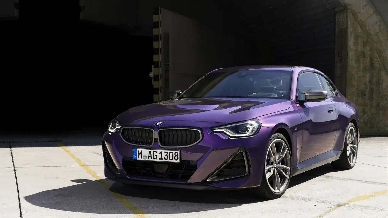 Fioletowe BMW 220i coupe z nowoczesnym designem, zaparkowane na betonowym placu przed ciemnym budynkiem.