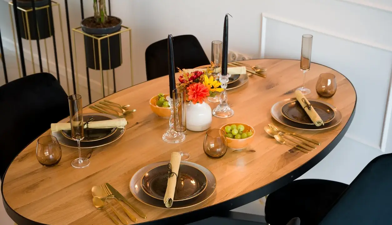 Un dîner élégant est prêt, avec des assiettes sombres, des couverts dorés et des raisins verts. Les art de la table sont mis en valeur par des bougies noires et des fleurs colorées.