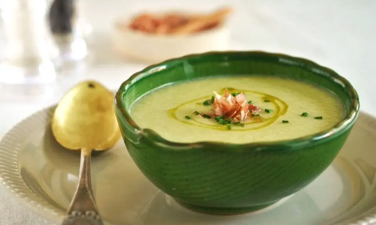 Un refrescante gazpacho de melón, adornado con jamón serrano y cebollino, servido en un cuenco verde con una cuchara dorada.