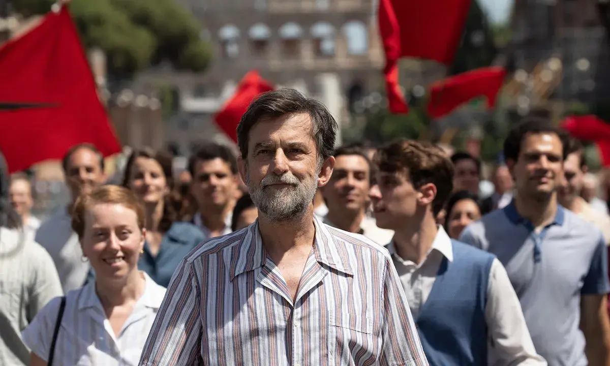 Nanni Moretti cammina in una folla con bandiere rosse, sullo sfondo il Colosseo.