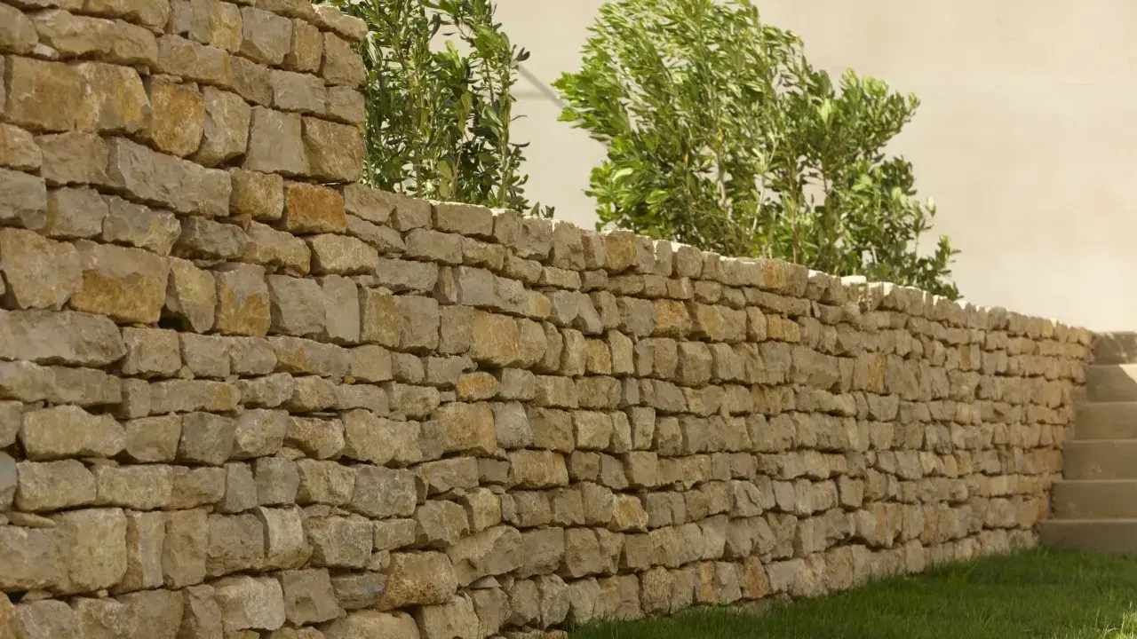 Un muret en pierre sèche, bordé d'herbe verte et de verdure luxuriante, s'étend sur la droite, menant à des marches en pierre.