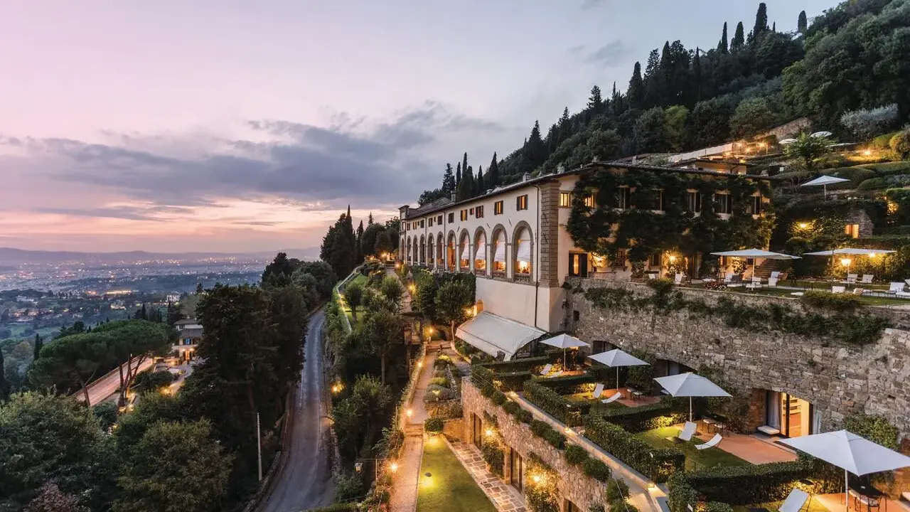 Un lussuoso hotel si affaccia sulle **colline di Firenze** al tramonto, con giardini illuminati e la città in lontananza.