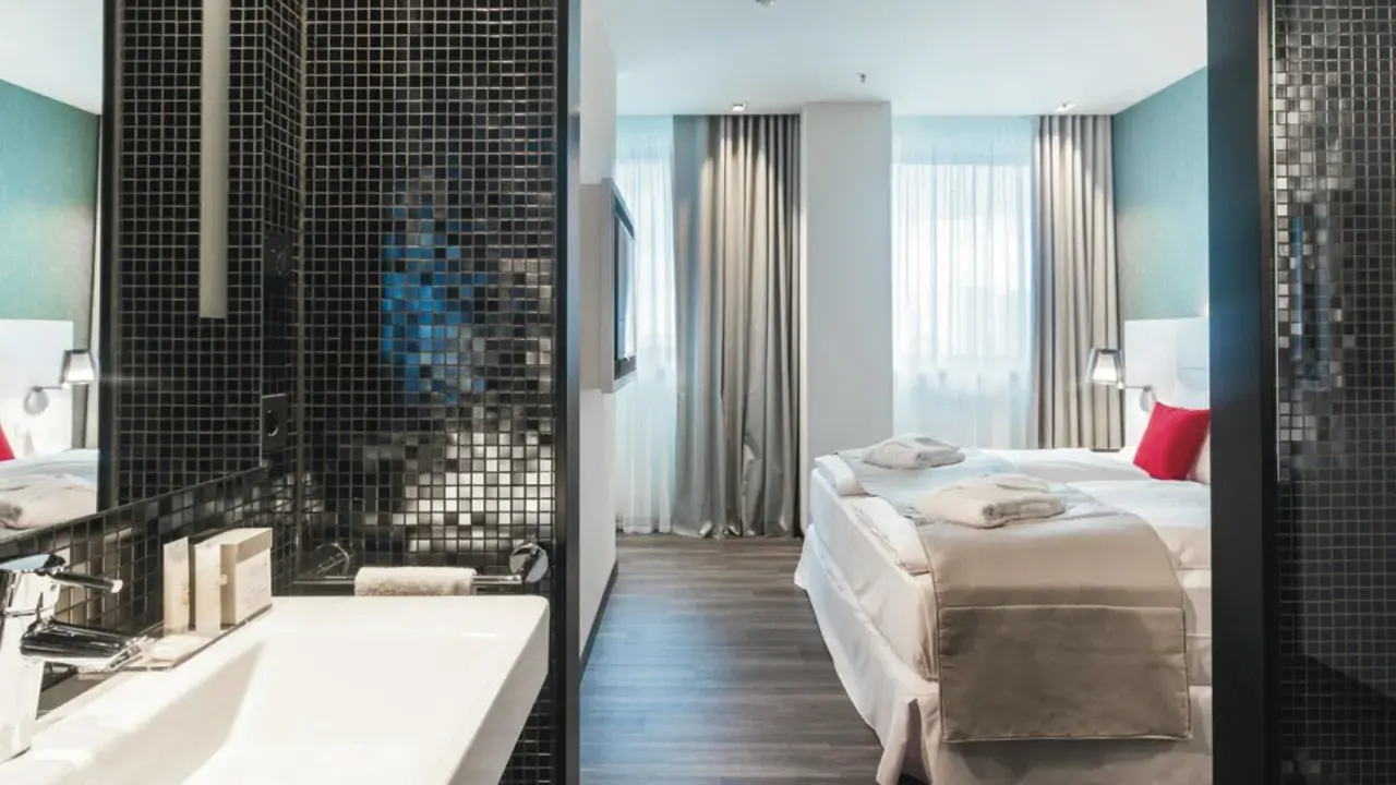 Badezimmer mit Waschbecken und Blick auf ein Hotelzimmer mit Bett und Fenstern.