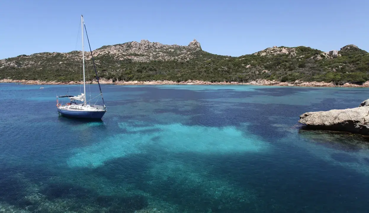 Un voilier bleu ancré dans les eaux turquoise de Cap Corse, avec des collines verdoyantes en arrière-plan sous un ciel clair.