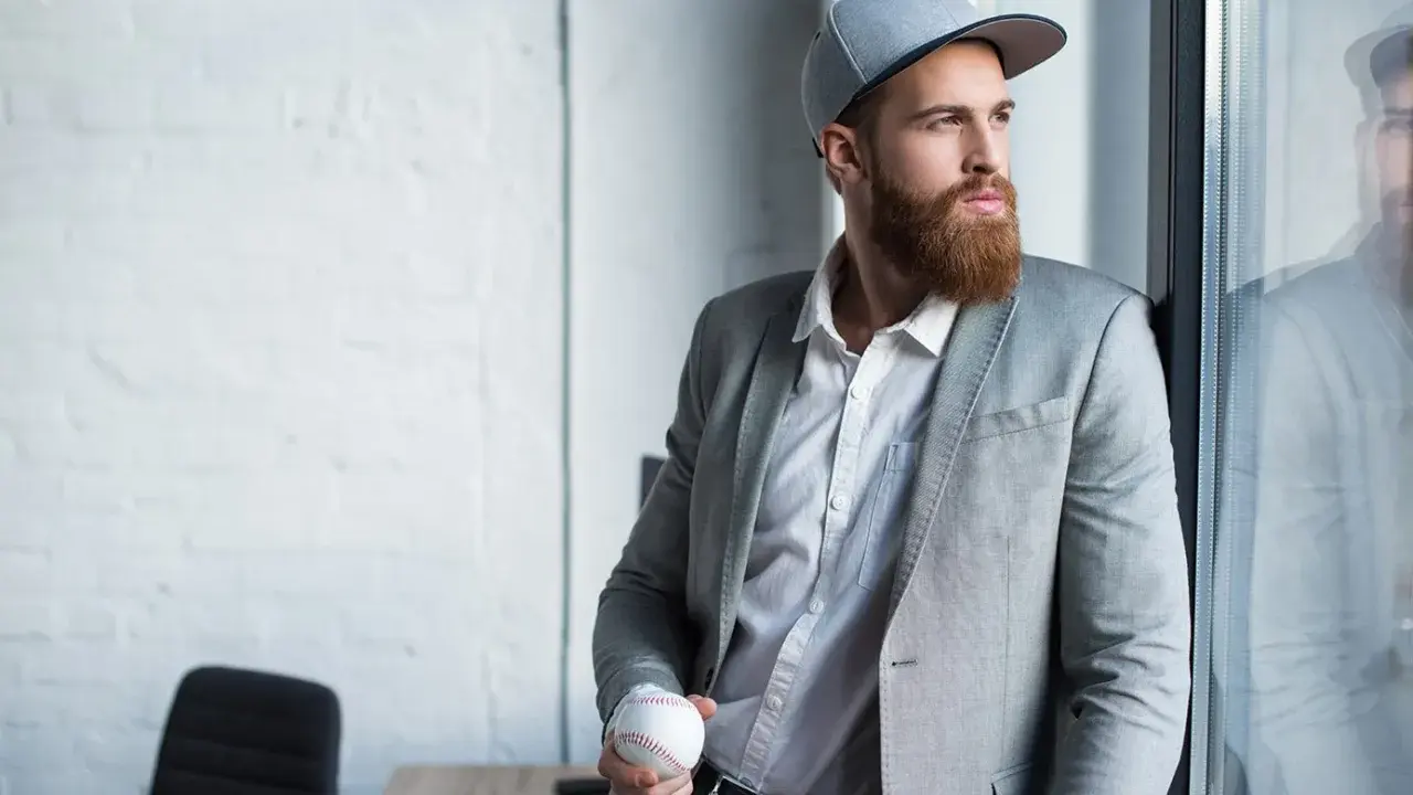 Mężczyzna w szarej czapce snapback i garniturze trzyma piłkę baseballową.