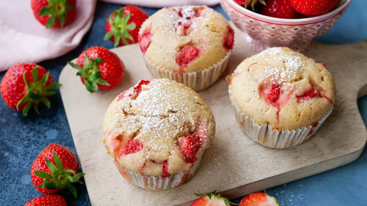Saftige Erdbeer Muffins, bestäubt mit Puderzucker, liegen auf einem Holzbrett, umgeben von frischen Erdbeeren.