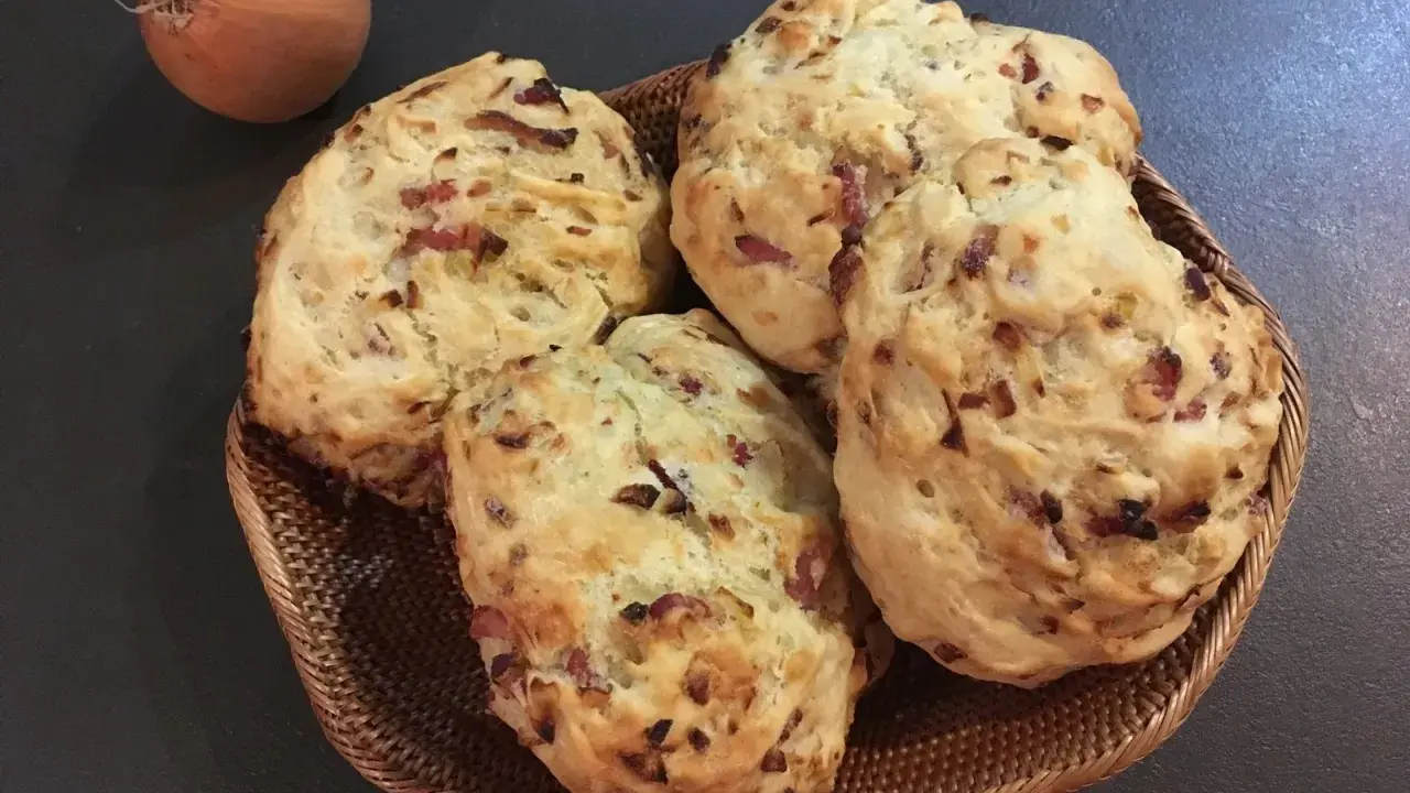 Quatre pains aux lardons et oignons dorés dans un panier en osier, avec un oignon cru à côté.