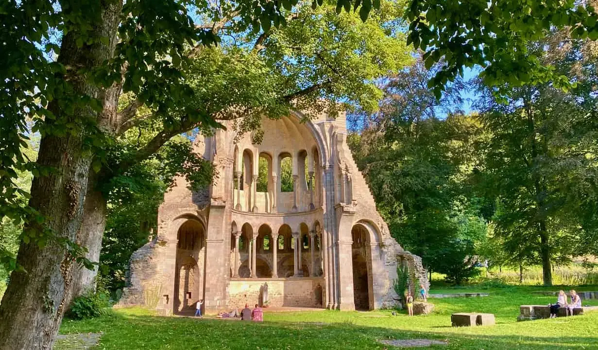 Ruinen einer alten Kirche in NRW, umgeben von Bäumen. Menschen genießen ihre Freizeit auf der grünen Wiese.