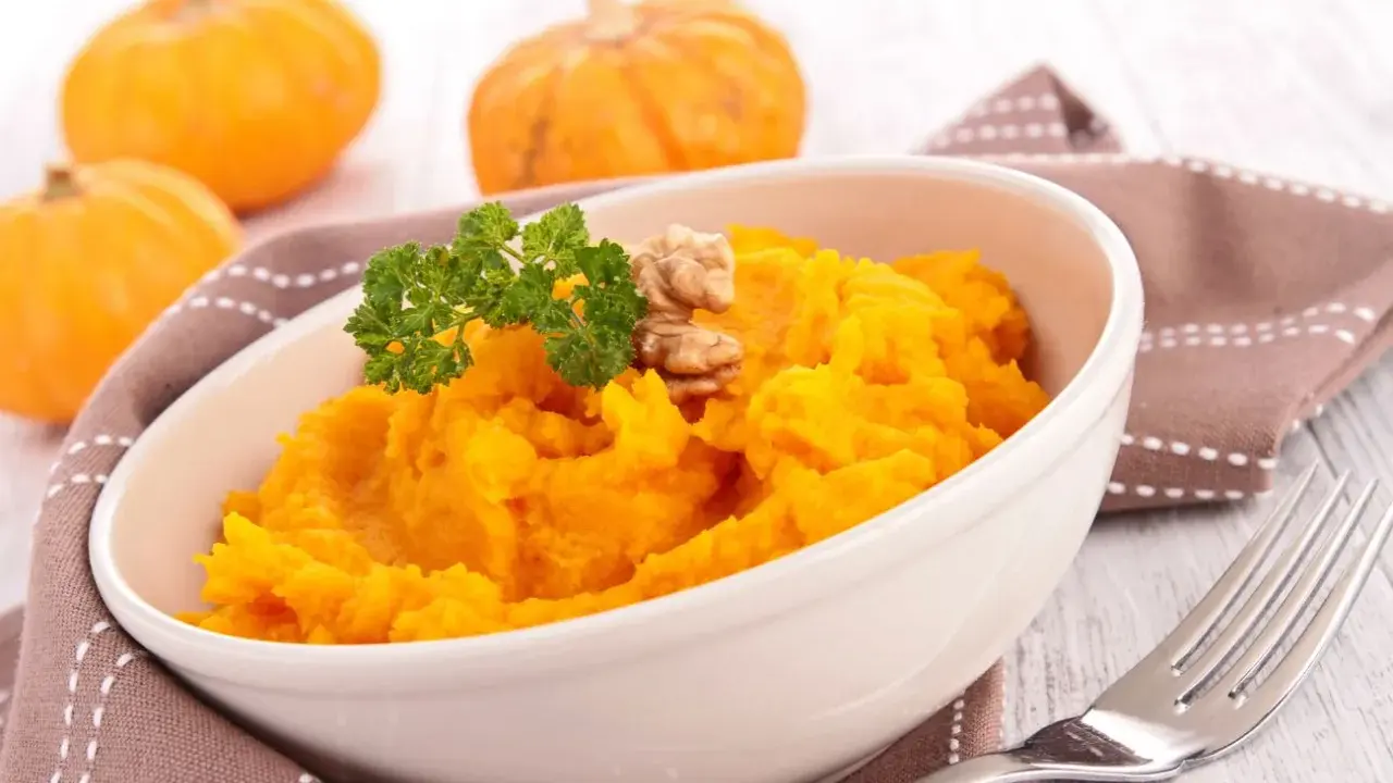 Un tazón de cremoso **puré de calabaza al horno**, adornado con perejil y nueces. Al fondo, calabazas pequeñas y una servilleta marrón.