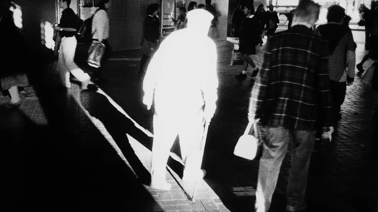 A stark black and white image by Trent Parke captures a blurred figure in a white hat and coat, walking with a cane. Other figures are also blurred, suggesting movement and a fleeting moment.