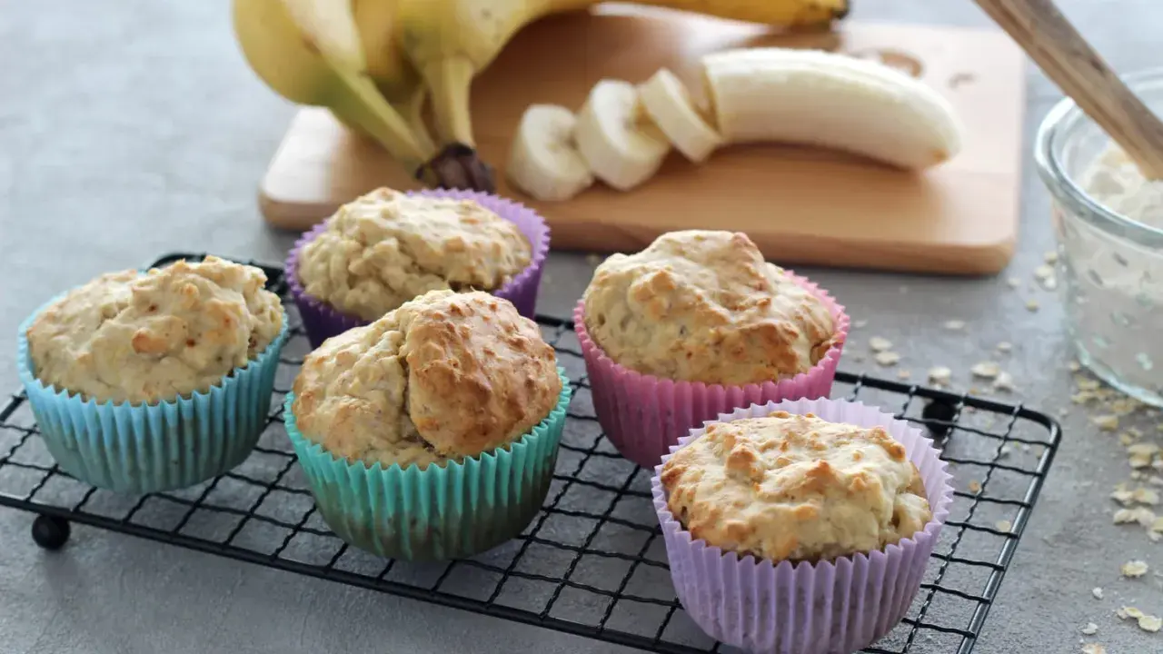 Fünf leckere bananenmuffins ohne zucker in bunten Förmchen auf einem Gitter. Bananen und Mehl im Hintergrund.