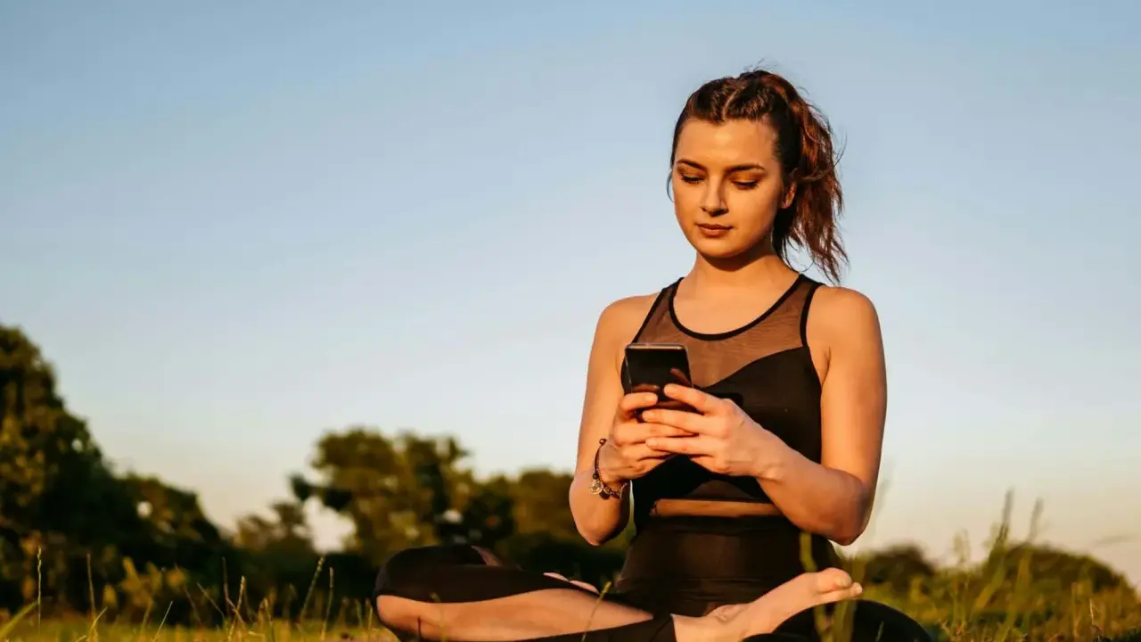 Kvinna i lotusställning på en matta utomhus, med en mobiltelefon i händerna. En modern typ av meditation.