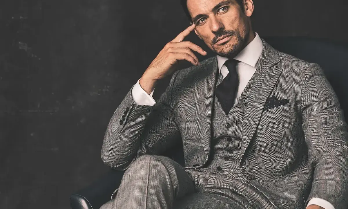 A man in a grey suit, black shirt, and tie, with a thoughtful expression, sits in a chair.