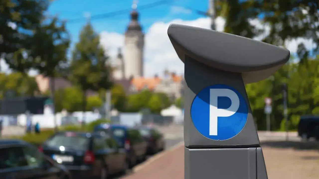 Parquímetro con señal de estacionamiento. Los coches eco pagan zona azul en esta calle urbana con edificios y árboles al fondo.