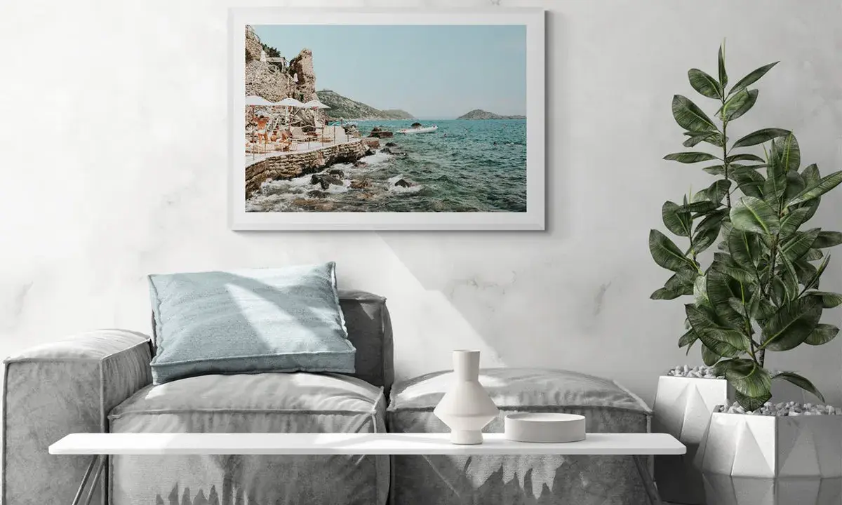 A framed print of a coastal scene, reminiscent of fine art photography galleries, hangs above a sofa. The artwork depicts a rocky shoreline with people relaxing under umbrellas.