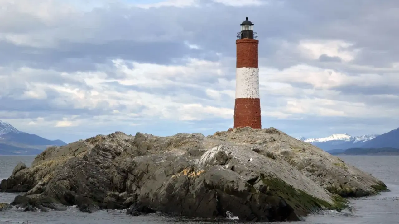 Faro a strisce rosse e bianche su un'isola rocciosa nel Canale di Beagle, con montagne innevate sullo sfondo.