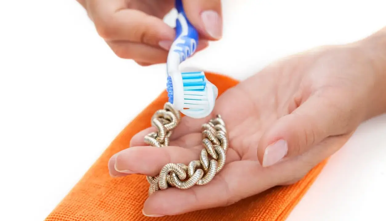 Manos limpian una pulsera de oro con un cepillo de dientes y un paño naranja. Aprende como limpiar el oro para que brille.