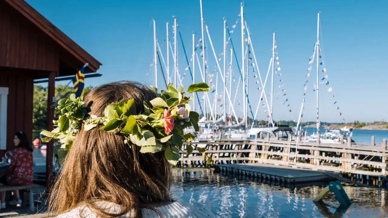 En flicka med blomsterkrans ser ut över en hamn med segelbåtar. En perfekt dag för en resa till Åland.