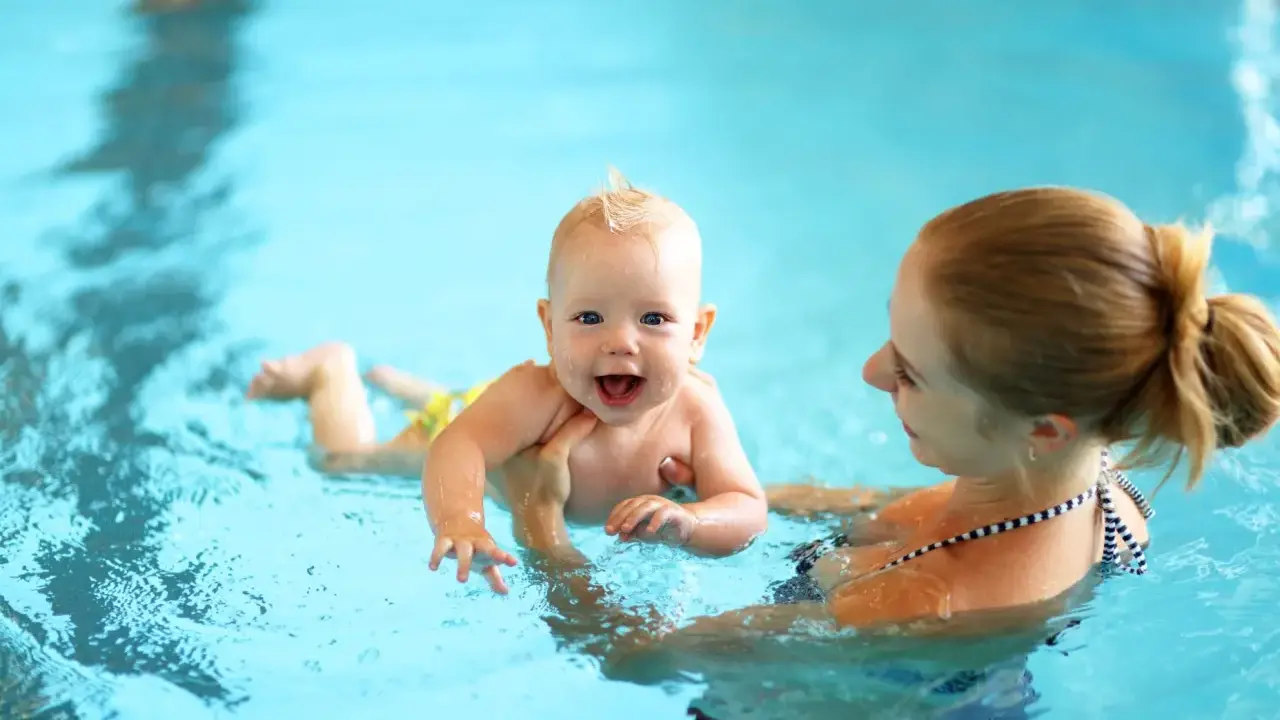 Glückliches 8 Wochen altes Baby mit Mutter im Schwimmbad. Das Baby lacht und planscht im Wasser, während die Mutter es festhält.