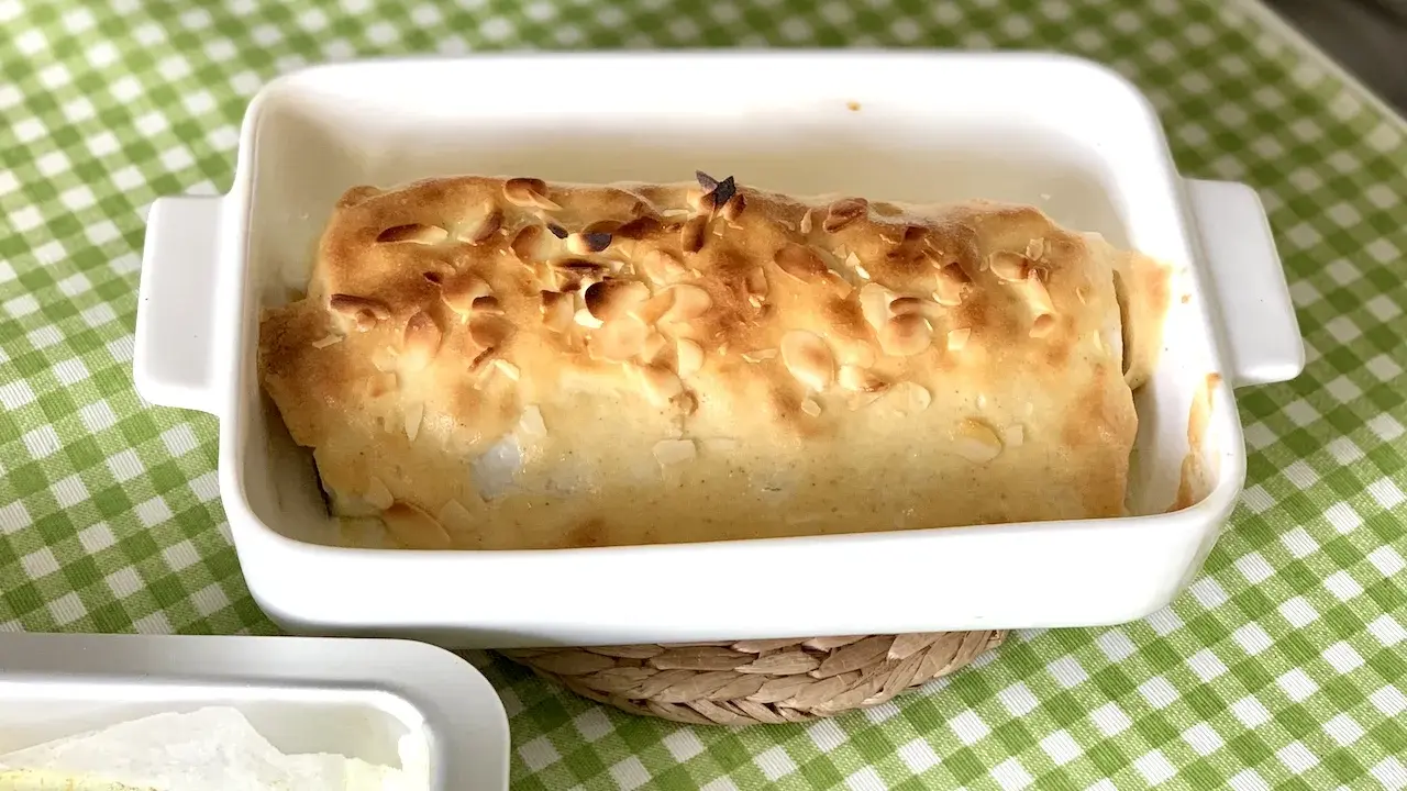 Bacalao gratinado con alioli, cubierto de almendras tostadas, en una fuente blanca sobre mantel de cuadros verdes.