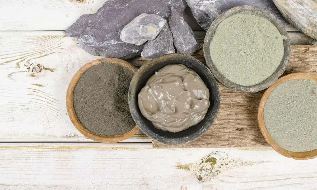 Pots de différentes argiles, dont une argile verte pour dégonfler, sur un fond en bois blanc et brut.