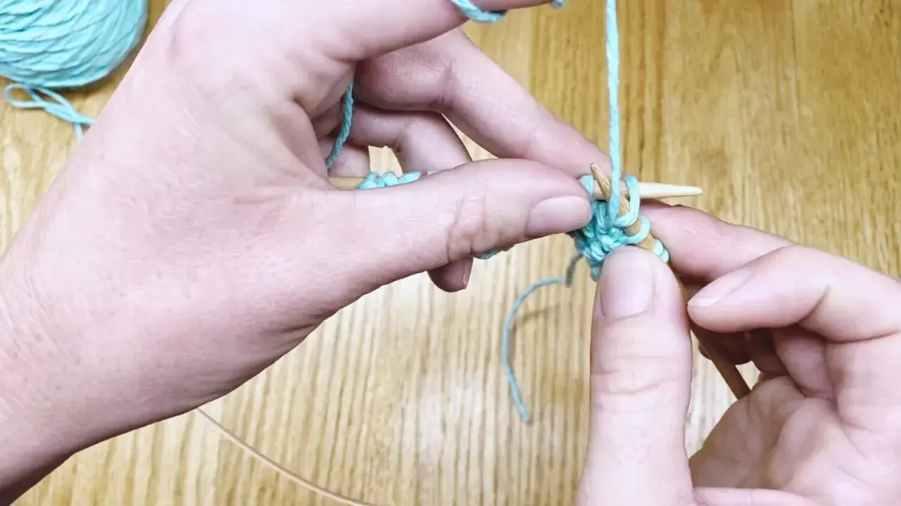 Manos tejiendo punto ojo de perdiz con hilo turquesa y agujas de madera.