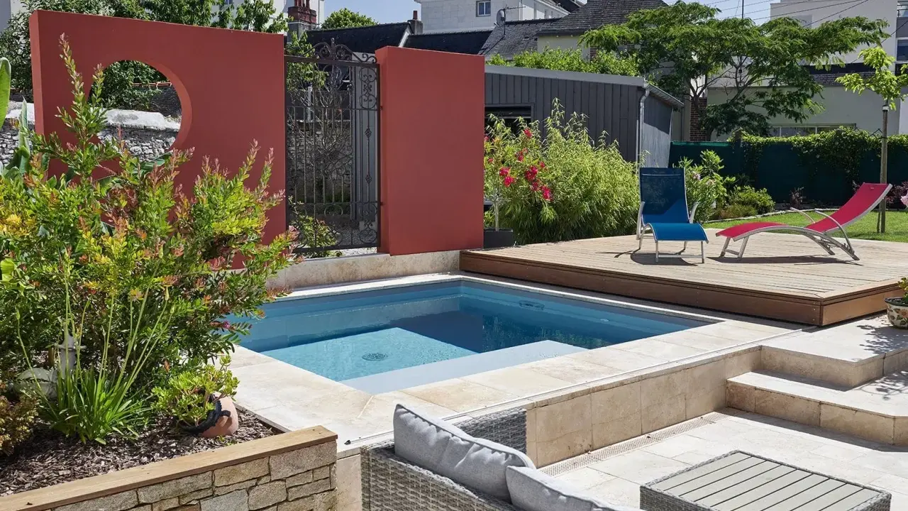 Une piscine moderne avec une belle décoration autour piscine : mur rouge, terrasse en bois, mobilier de jardin et végétation luxuriante.
