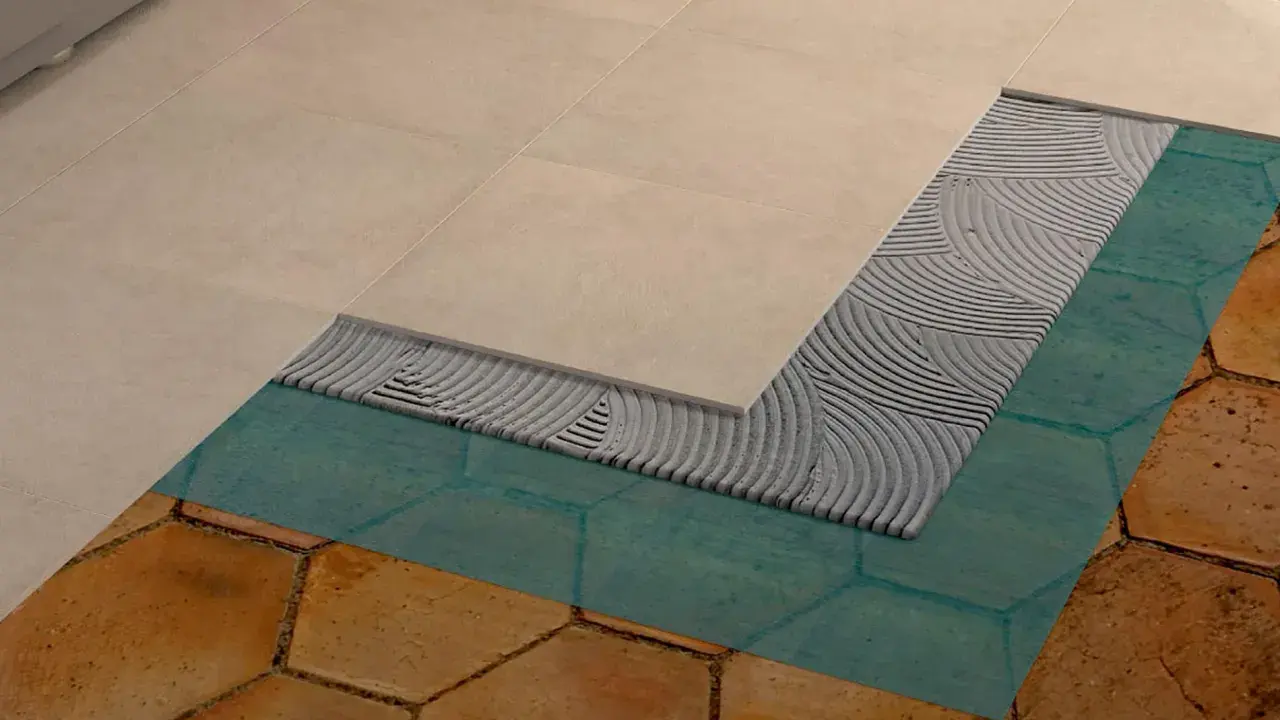Dettaglio di un pavimento in fase di posa: nuove piastrelle beige sopra uno strato di colla, membrana impermeabilizzante turchese e vecchie piastrelle esagonali color terra.