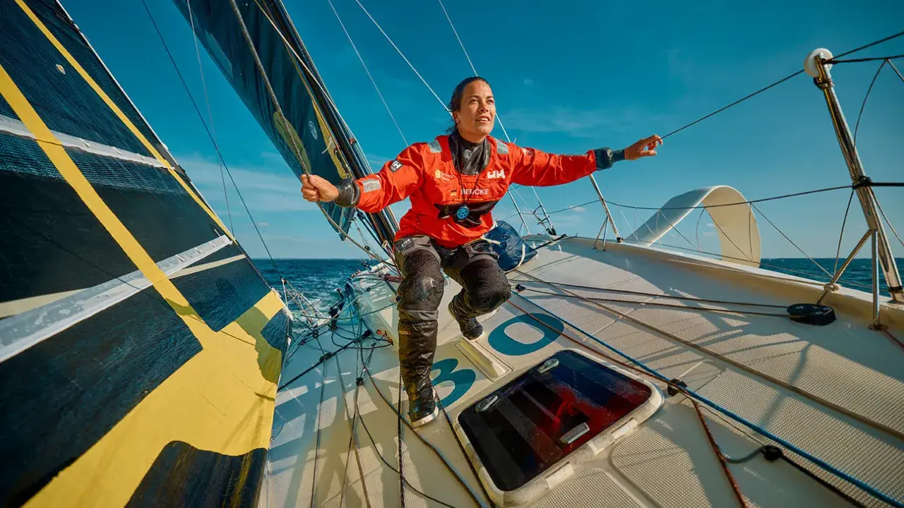 Sanni Beucke steuert ihr Segelboot mit voller Kraft über das Meer. Sie trägt eine orangefarbene Jacke und blickt entschlossen nach vorn.