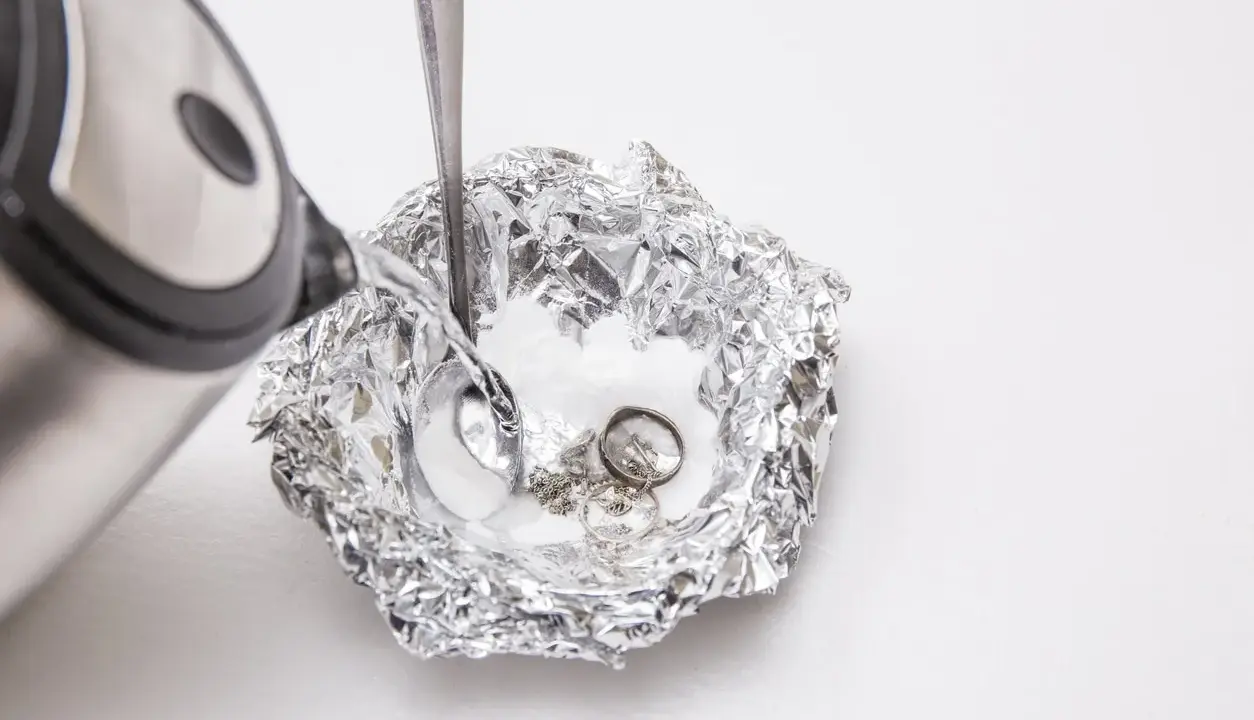Nettoyage d'argenterie noircie : de l'eau chaude est versée sur des bijoux en argent et une cuillère dans une coupelle en papier aluminium.